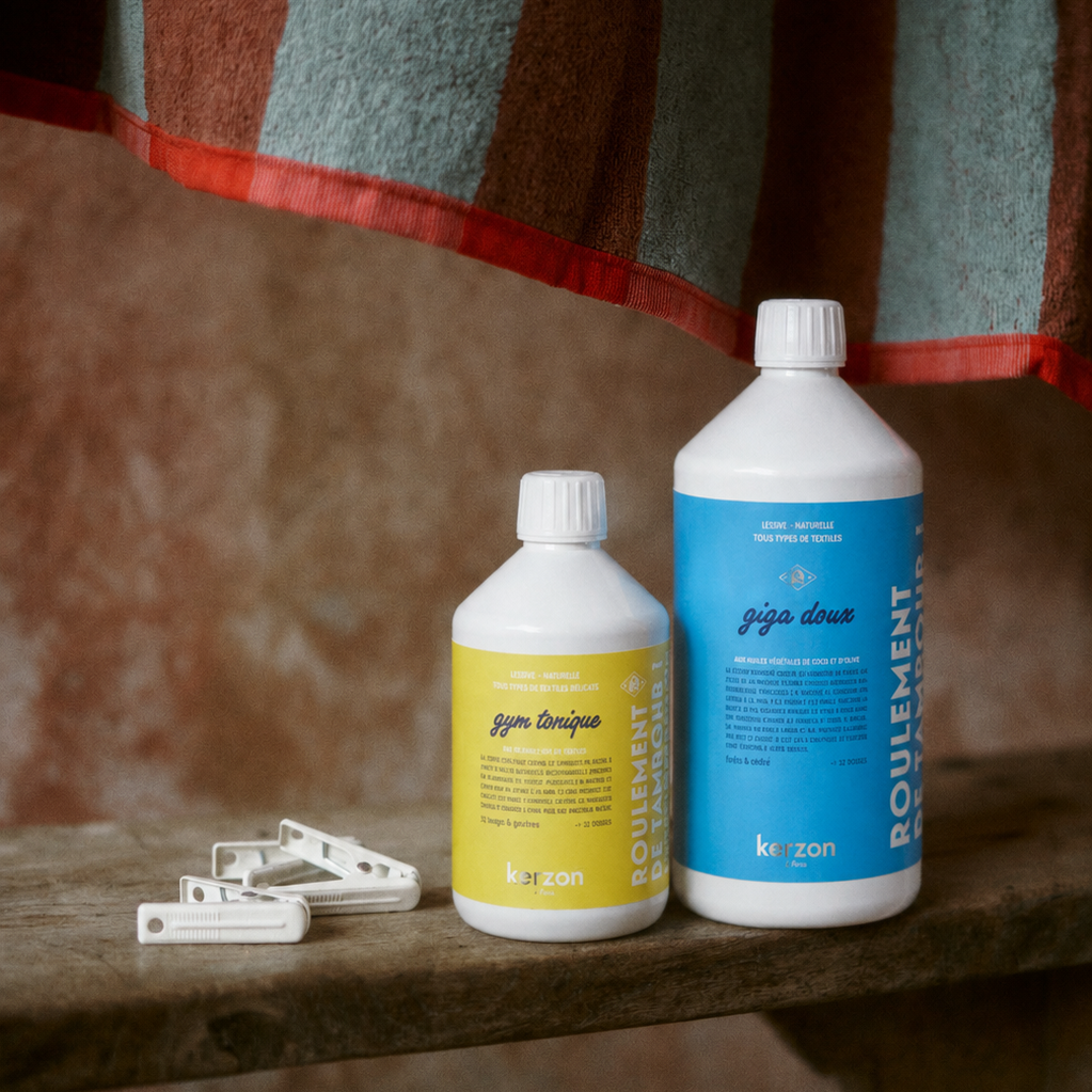 Two bottles of Kerzon laundry soap, one with a yellow label and one with a blue label, displayed on a rustic wooden shelf with white clothespins in the foreground and a striped towel hanging in the background.