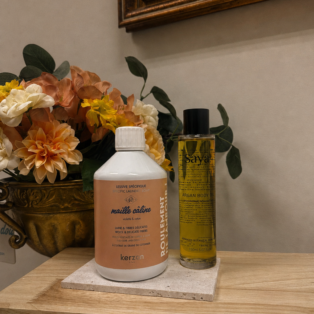 laundry wash from Kerzon and body oil from Saya displayed on a marble coaster on a wooden shelf, with a vase of flowers and a framed picture in the background.