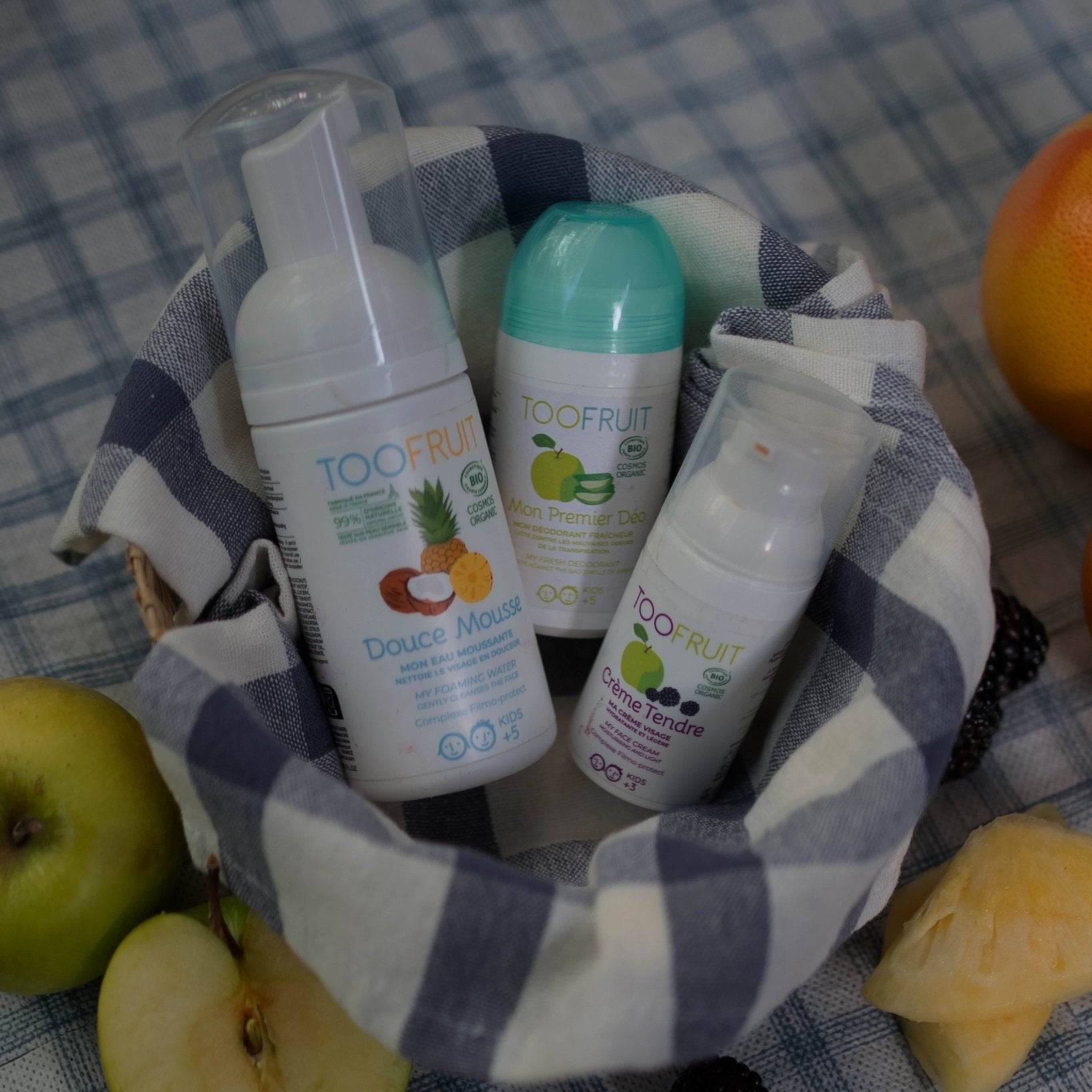 Three 'TOOFRUIT' skincare products in a basket with fruits on a checkered tablecloth